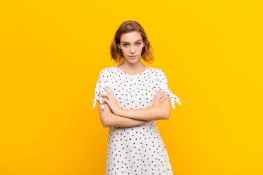 Genç sarışın kadın hayal kırıklığına uğramış, hayal kırıklığına uğramış, ciddi görünüyor, kızgın ve kollarını düz duvara karşı çapraz yapmış.