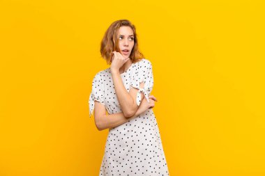 Genç sarışın kadın düşünüyor, kuşkulu ve şaşkın hissediyor, farklı seçenekleri var, düz renkli duvara karşı hangi kararı vereceğimi merak ediyor.