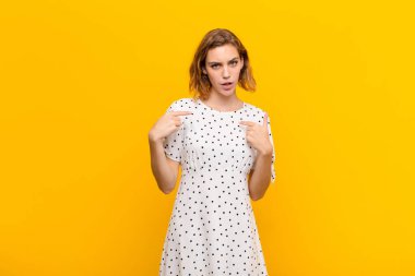 Genç sarışın kadın, kafası karışmış ve tuhaf bakışlarıyla kendini işaret ediyor. Düz renkli duvara karşı seçildiği için şaşırmış ve şaşırmış.