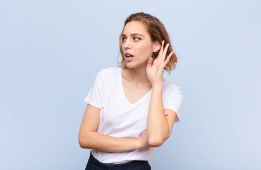 Genç sarışın kadın ciddi ve meraklı görünüyor, dinliyor, gizli bir konuşma ya da dedikodu duymaya çalışıyor, düz renkli duvara kulak misafiri oluyor.