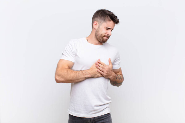 молодой ручной человек смотрит грустно, больно и с разбитым сердцем, держа обе руки близко к сердцу, плача и чувствуя депрессию против плоской стены цвета
