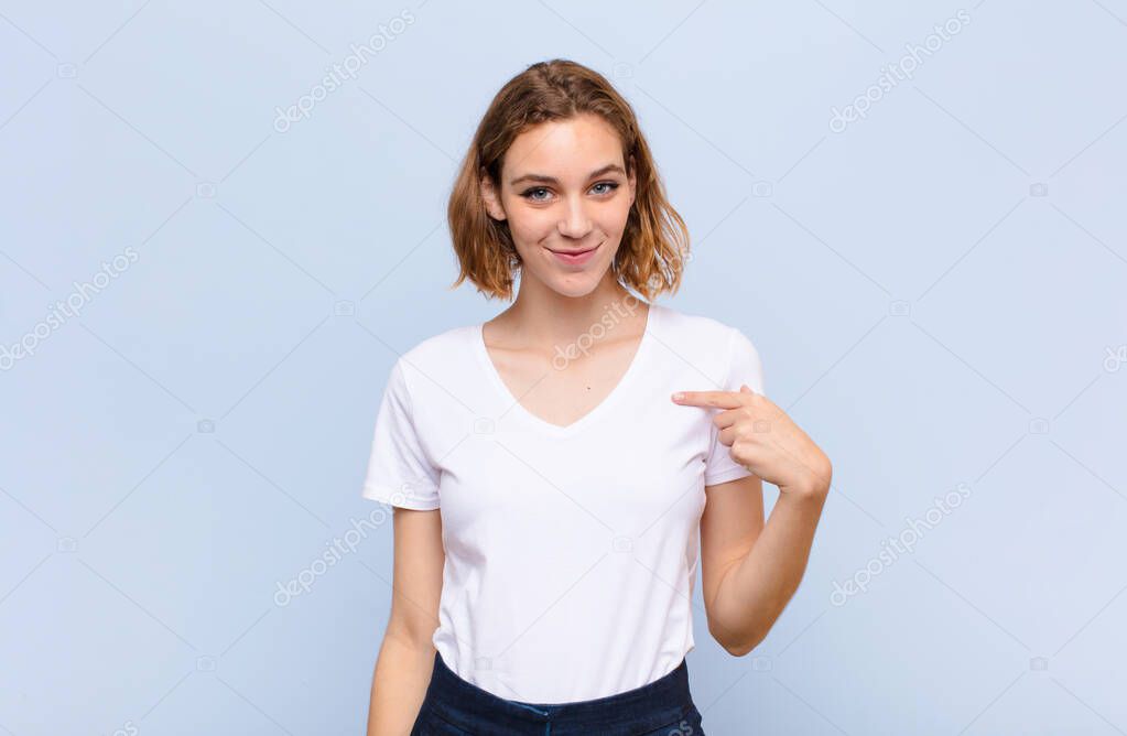 young blonde woman looking proud, confident and happy, smiling and pointing to self or making number one sign against flat color wall