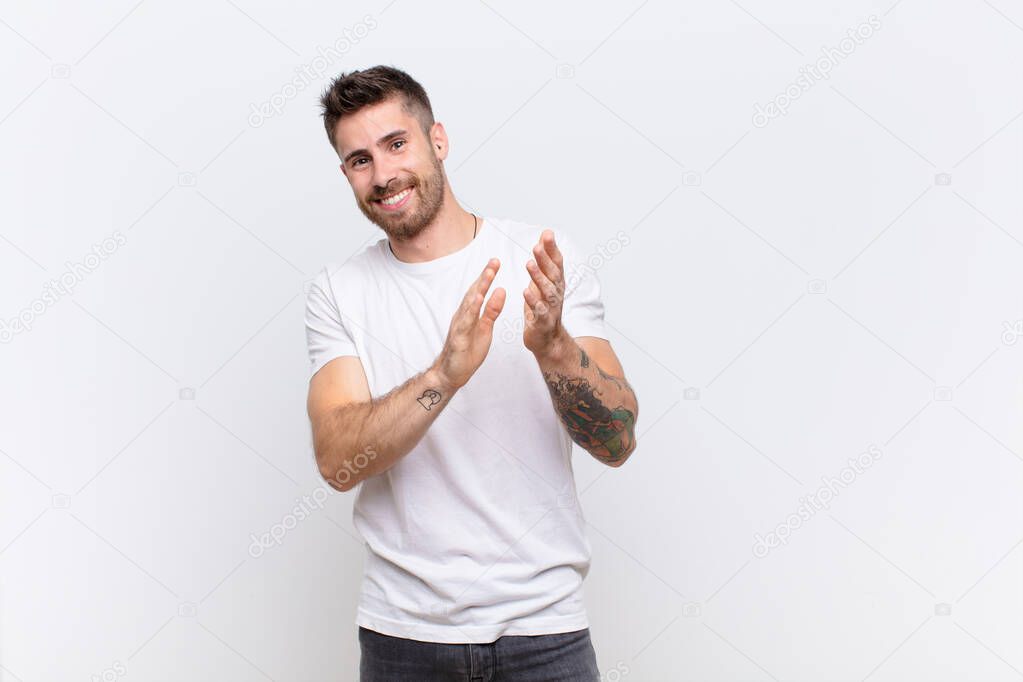 young handosme man feeling happy and successful, smiling and clapping hands, saying congratulations with an applause against flat color wall