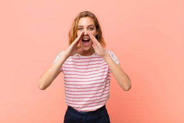 Genç sarışın kadın mutlu, heyecanlı ve pozitif hissediyor. Elleri ağızda, düz duvara karşı bağırıyor.