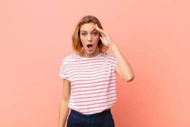 Genç sarışın kadın mutlu, şaşkın ve şaşırmış görünüyor, gülümsüyor ve düz renkli duvara karşı inanılmaz ve inanılmaz iyi haberler veriyor.