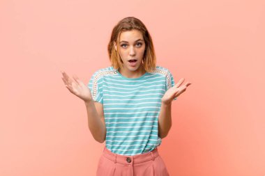 Genç sarışın kadın şok olmuş, şaşırmış, endişeli ve paniklemiş hissediyor. Düz renkli duvara karşı stresli ve dehşetli bir bakışla bakıyor.