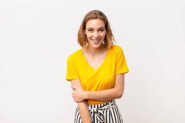 Genç sarışın kadın utangaç ve neşeli bir şekilde gülüyor, arkadaş canlısı ve olumlu ama düz renkli duvara karşı güvensiz bir tutum sergiliyor.