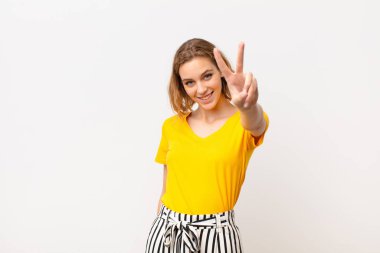 Genç sarışın kadın gülümsüyor ve mutlu görünüyor, kaygısız ve pozitif, bir eliyle düz duvara karşı zafer ya da barış işareti yapıyor.