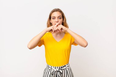 Genç sarışın kadın ciddi ve hoşnutsuz görünüyor. Reddedilmek için iki parmağını öne uzatıyor. Düz renkli duvara karşı sessizlik istiyor.