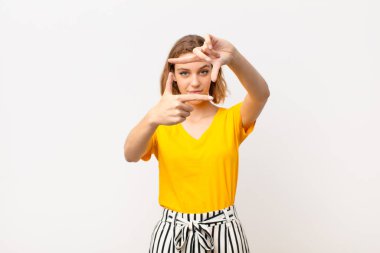 Genç sarışın kadın mutlu, arkadaş canlısı ve pozitif hissediyor, gülümsüyor ve düz renkli duvara karşı elleri ve fotoğraf çerçevesi yapıyor.