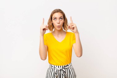 Genç sarışın kadın şaşırmış, şaşırmış ve ağzı açık görünüyor, iki eliyle yukarıya doğru bakıyor düz renkli duvara uzayı kopyalamak için.