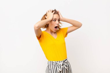 Korkunç bir hata yüzünden dehşete düşmüş ve şok olmuş görünen ağzı açık genç sarışın kadın düz renkli duvara karşı el kaldırıyor.