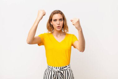 Genç sarışın kadın inanılmaz bir başarıyı kutluyor. Kazanan gibi, heyecanlı ve mutlu görünüyor. Al bakalım! yassı renk duvarına karşı