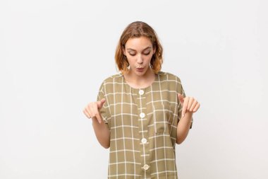 Genç sarışın kadın, ağzı açık, iki eliyle aşağıya bakıyor, şaşırmış, şaşırmış ve şaşırmış görünüyor.