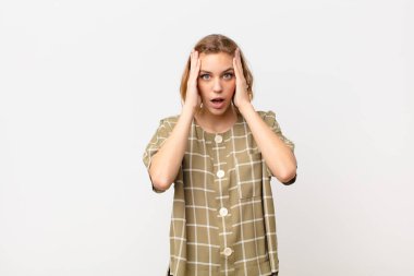 Genç sarışın kadın hoş olmayan bir şekilde şok olmuş, korkmuş ya da endişeli görünüyor, ağzı sonuna kadar açık ve iki kulağını da düz renkli duvara karşı eliyle kapatıyor.