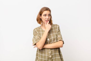 Genç sarışın kadın düşünüyor, kuşkulu ve şaşkın hissediyor, farklı seçenekleri var, düz renkli duvara karşı hangi kararı vereceğimi merak ediyor.