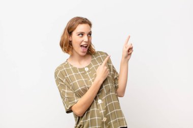 Genç sarışın kadın sevinçli ve şaşırmış hissediyor, şok olmuş bir ifadeyle gülümsüyor ve düz renkli duvara işaret ediyor.