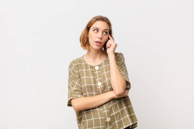 Yoğunlaşmış bakışlı genç sarışın kadın, şüpheli bir ifadeyle, düz renkli duvara bakıp duruyor.