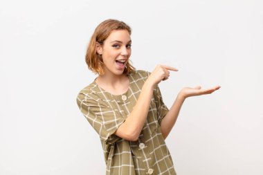 Genç sarışın kadın neşeyle gülümsüyor ve yan taraftaki fotokopi alanına işaret ediyor, düz renkli duvara karşı bir nesneyi gösteriyor veya reklamını yapıyor.