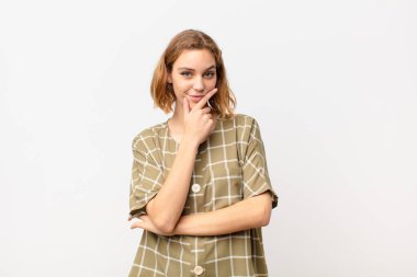 Genç sarışın kadın ciddi, düşünceli ve güvensiz görünüyor, bir kolu çapraz ve çene üzerinde, düz renkli duvarla seçenekleri tartıyor