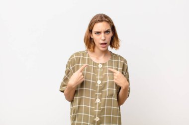 Genç sarışın kadın, kafası karışmış ve tuhaf bakışlarıyla kendini işaret ediyor. Düz renkli duvara karşı seçildiği için şaşırmış ve şaşırmış.