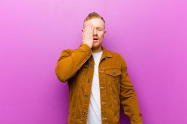 Genç sarışın adam uykulu, sıkılmış ve esniyor, baş ağrısı ve yüzünün yarısını mor arka planla kaplayan bir el