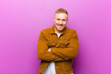 Genç sarışın adam mutlu, gururlu ve memnun görünüyor. Kolları mor arka planda çaprazlanmış başarılı bir şekilde gülümsüyor.