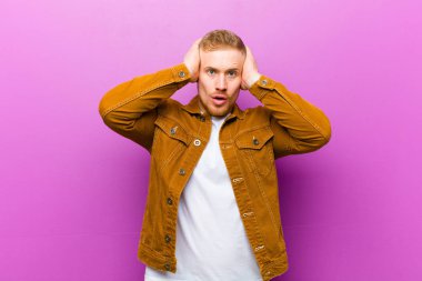 Genç sarışın adam heyecanlı ve şaşırmış görünüyor, ağzı açık, iki eli başının üstünde, mor arka planda şanslı bir kazanan gibi hissediyor.