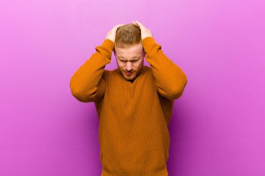 Kazak giyen genç sarışın bir adam stresli ve hayal kırıklığına uğramış hissediyor, ellerinizi havaya kaldırıyor, yorgun, mutsuz ve migren ağrısı çekiyor.