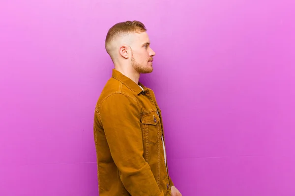 young blonde man on profile view looking to copy space ahead, thinking, imagining or daydreaming against purple background