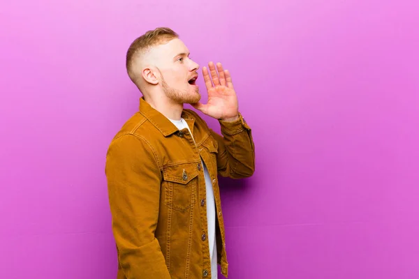 young blonde man profile view, looking happy and excited, shouting and calling to copy space on the side against purple background