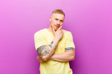 Sarı tişörtlü genç bir adam ciddi, düşünceli ve güvensiz görünüyor. Bir kolu çapraz ve eli çenesinde, seçenekleri tartıyor.