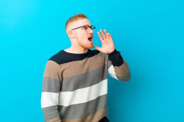 young blonde man yelling loudly and angrily to copy space on the side, with hand next to mouth against blue background