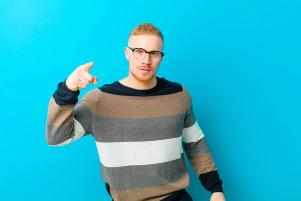 young blonde man pointing at camera with an angry aggressive expression looking like a furious, crazy boss against blue background