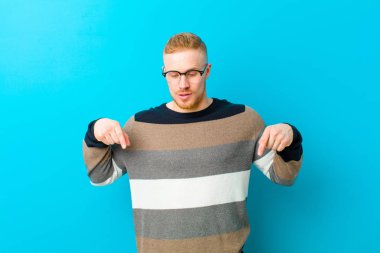young blonde man with open mouth pointing downwards with both hands, looking shocked, amazed and surprised against blue background