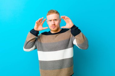 young blonde man screaming with hands up in the air, feeling furious, frustrated, stressed and upset against blue background