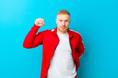 Genç sarışın adam spor kıyafetleri giyiyor ciddi, güçlü ve asi hissediyor, yumruğunu kaldırıyor, protesto ediyor ya da mavi arka plana karşı devrim için savaşıyor.