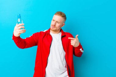 Mavi arka planda spor giysileri giyen, su şişeli genç sarışın bir adam. fitness konsepti