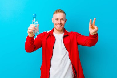 Mavi arka planda spor giysileri giyen, su şişeli genç sarışın bir adam. fitness konsepti