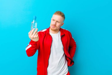 Mavi arka planda spor giysileri giyen, su şişeli genç sarışın bir adam. fitness konsepti