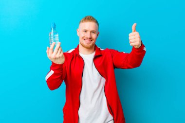 Mavi arka planda spor giysileri giyen, su şişeli genç sarışın bir adam. fitness konsepti