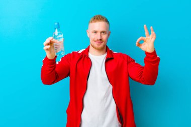 Mavi arka planda spor giysileri giyen, su şişeli genç sarışın bir adam. fitness konsepti