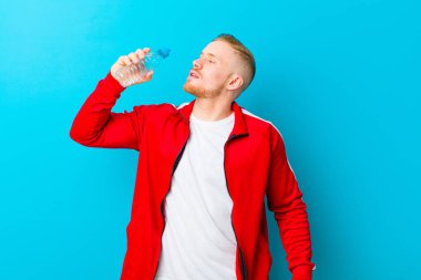 Mavi arka planda spor giysileri giyen, su şişeli genç sarışın bir adam. fitness konsepti