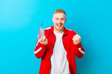 Mavi arka planda spor giysileri giyen, su şişeli genç sarışın bir adam. fitness konsepti