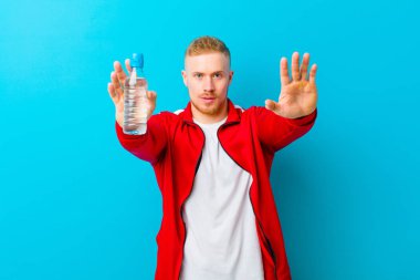 Mavi arka planda spor giysileri giyen, su şişeli genç sarışın bir adam. fitness konsepti