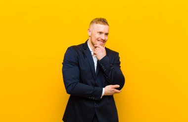 Genç sarışın iş adamı mutlu ve kendinden emin bir ifadeyle gülümsüyor. Elleri çenesinde, merak ediyor ve yan tarafa bakıyor.