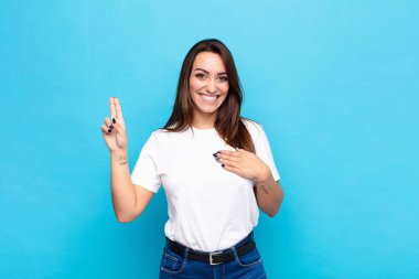 Genç, güzel bir kadın mutlu, kendinden emin ve güvenilir görünüyor. Gülümsüyor ve zafer işareti gösteriyor. Mavi duvara karşı olumlu bir tutum sergiliyor.