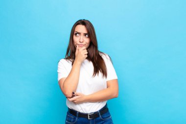 Genç ve güzel bir kadın düşünceli, meraklı ya da hayal kuruyor, hayal kuruyor ve mavi duvara uzayı kopyalamak için yukarı bakıyor.