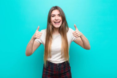 Genç ve güzel bir kadın gülümsüyor. Mutlu, pozitif, kendinden emin ve başarılı görünüyor. İki başparmağı da mavi duvarda.