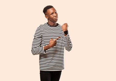 Genç Afro-Amerikan siyahi adam sabırsız ve kızgın görünüyor, saati gösteriyor, dakiklik istiyor, bej duvara karşı zamanında olmak istiyor.